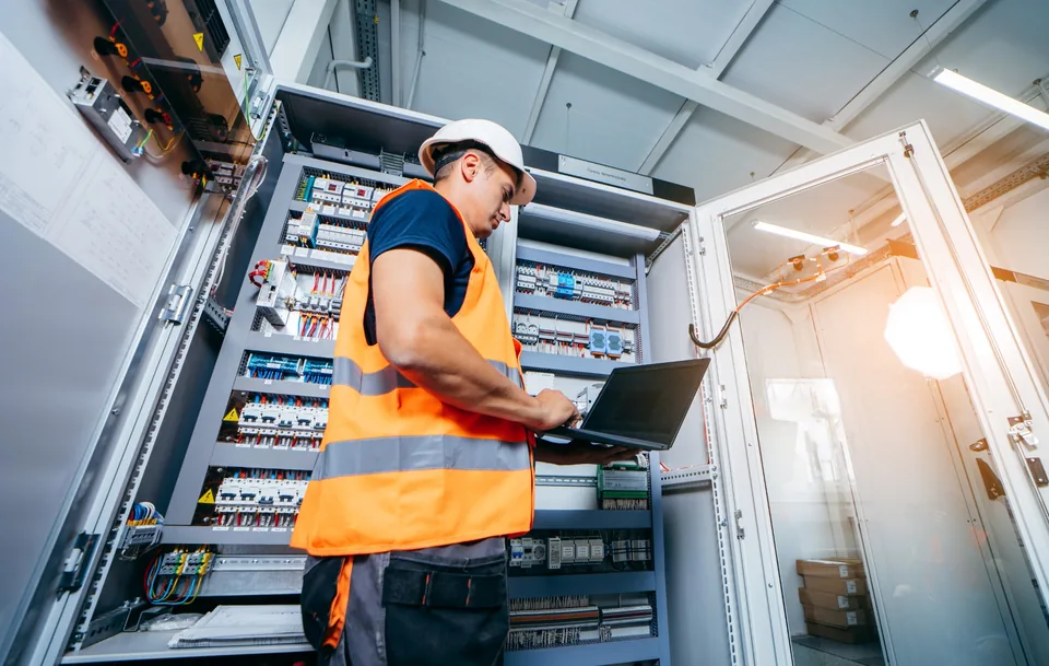 Industrial electrical technician using a laptop in a control room for planned electrical maintenance and system assessment